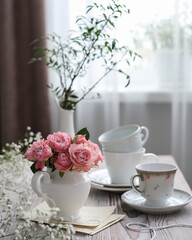 Fototapeta na wymiar A vase of pink roses and white cups on a table by the window. Spring, summer still life