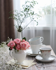 A vase of pink roses and white cups on a table by the window. Spring, summer still life
