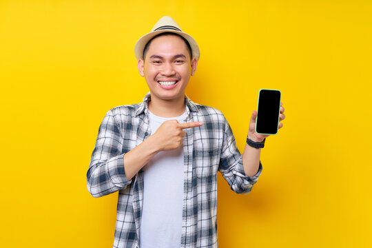 Smiling Handsome Asian Young Man 20s Wearing Casual Clothes Hat Pointing Finger At Mobile Phone With Blank Screen Isolated On Yellow Background. People Lifestyle Concept