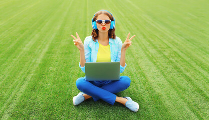 Happy young woman with laptop listening to music in headphones in the park on green grass background