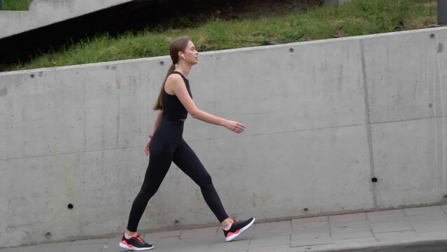 Young sporty woman with earphones walking to rest after running outdoors, camera moving right