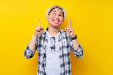 Smiling Young handsome ethnic Asian man 20s wearing casual clothes hat pointing finger overhead indicating on workspace area copy space mockup isolated on yellow background. People lifestyle concept