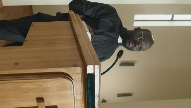 Vertical medium long shot of young adult Black priest standing at wooden lectern with microphone on it preaching to parishioners