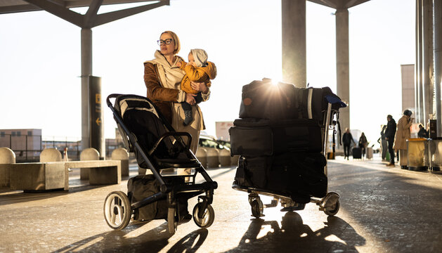 Motherat travelling with his infant baby boy child, walking, pushing baby stroller and luggage cart in front of airport terminal station