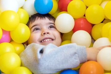 Fototapeta na wymiar kid having fun in play center pool with colorful many balls.happy smiling child preschooler boy throwing tossing up plastic balls playground interior inside mall.soft ocean plastic balls cover face