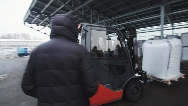Warehouse loader moves polycarbonate building materials by electric forklift. Worker transports package on wooden pallet to storage place under lean-to