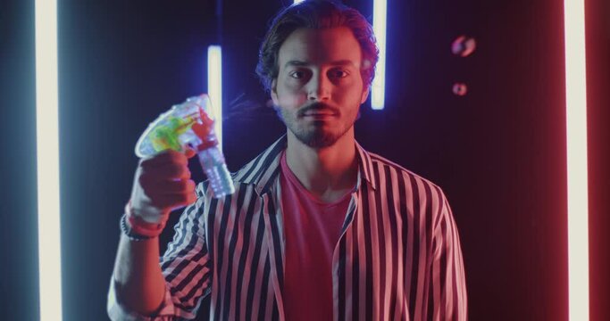 Portrait of serious Middle Eastern man shooting toy gun with soap bubbles on futuristic neon background. People and entertainment for adult concept.