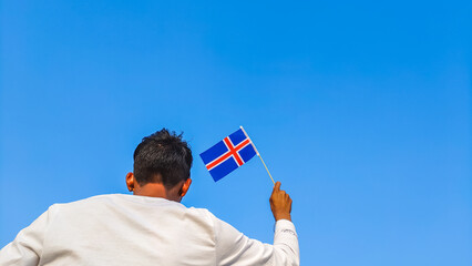 Boy holding Iceland flag against clear blue sky. Man hand waving Icelandic flag view from back, copy space