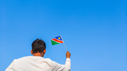 Boy holding Namibia flag against clear blue sky. Man hand waving Namibian flag view from back, copy...