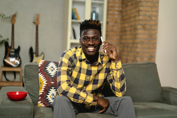 Handsome African man using smartphone while sitting on a sofa in cozy living room. Young man talking to the phone