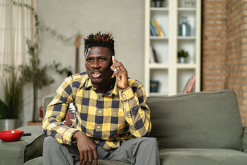Handsome African man using smartphone while sitting on a sofa in cozy living room. Young man talking to the phone