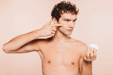 Fototapeta premium Close up portrait of handsome young man with healthy skin, applies cream moisturizer for anti wrinkle or anti aging, cares of body, poses naked or shirtless, isolated over beige background.