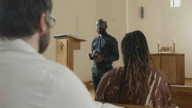 Selective focus shot of modern African American priest saying sermons to people in Catholic church