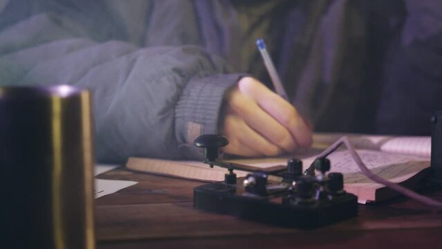 Man sends signal in Morse code tapping finger and writing notes in notebook. Male person sits at wooden table in dark room close view