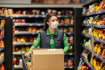 A stock clerk with face mask is pushing shopping cart full of products and filling up aisles in supermarket during covid.