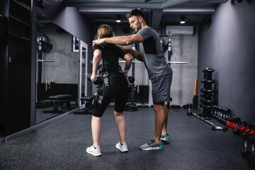 Fototapeta na wymiar A young male personal trainer assists a woman in an electric muscle suit to stimulate muscles. Fitness in an EMS suit in a modern gym concept, revolutionary training and care, electrical stimulation