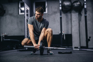 Naklejka na ściany i meble Preparing for strong muscle burning training. A young attractive man in gray sportswear sets up barbell weights in the gym. Fit young man looking focused on practice, sports discipline