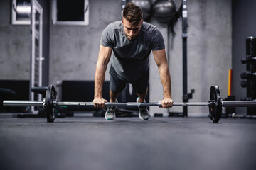 Fototapeta na wymiar Good physical shape of a sporty male person. A man in a plank position keeps his hands on a barbell and does push-ups in an indoor gym with a gray interior. Fitness goal, indoors workout