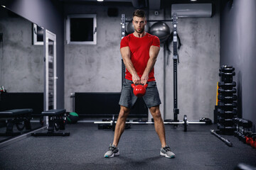 Fototapeta na wymiar A confident attitude and a seductive look of a muscular sportsman performing exercises with a kettle bell. Dead lifts and squats with a load in the indoor space of a modern gym.