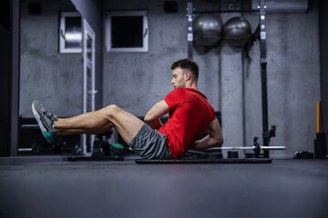 Obraz na płótnie Canvas Strengthening the body core and abdominal muscles. A sporty young guy does sit-ups on the side in a sporty red shirt and gray shorts on the floor of a modern gym. Sports routine, healthy living