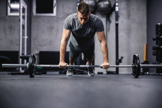Good physical shape of a sporty male person. A man in a plank position keeps his hands on a barbell and does push-ups in an indoor gym with a gray interior. Fitness goal, indoors workout