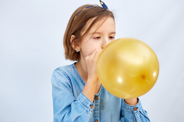 Pretty little girl in casual denim dress blowing, inflate yellow balloon on white background, studio