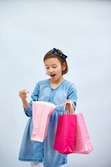 Pretty child girl in casual denim dress hold in hand pink shopping bags on white background, studio, Shopaholic shopping and fashion, discount sale concept, copy space, banner. Black friday, WOW