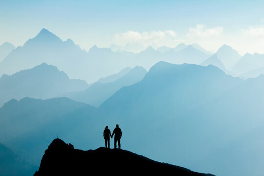 Silhouette Couple of man and woman reaching mountain top enjoying freedom and looking towards blue mountain silhouettes and sunrise. Alps, Allgaeu, Bavaria, Germany.
