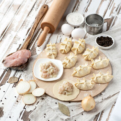 Manti on wooden cutting board, central asian cuisine. Manti making process. Minced chicken meat filling, ingredients for dough, spices, herbs and kitchen tools on wooden table. Ready-made semi