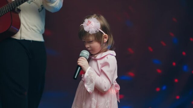 Little girl on stage in vintage dress, she sings into microphone and dances, her father plays acoustic guitar. Color music is shining. Performance on stage. Early development of children
