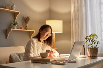 Portrait of serious concentrated woman with brown hair sitting at table working on laptop in evening at home, writing and working online, spending time with learning,