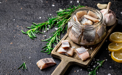 Salted herring in a jar on a cutting board with rosemary and lemon. 