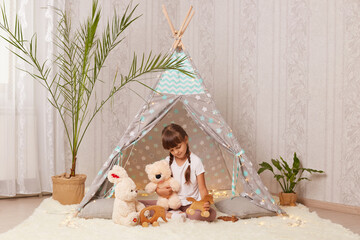 Photo of smiling calm little girl with pigtails wearing white t-shirt playing with toys in wigwam, sitting on floor on soft carpet, holding fluffy teddy bear and wooden horse. © sementsova321