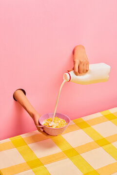 Food pop art photography. Female hands sticking out pink paper, pouring milk into bowl with corn flakes, ceral. Breakfast. Taste, creativity, art. Complementary colors. Copy space for ad, text