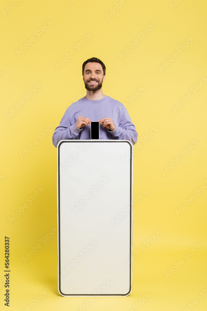 Poster bearded man smiling at camera and showing smartphone with blank screen near white phone template on yellow background.