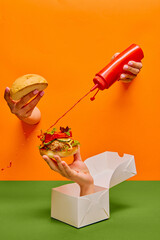 Food pop art photography. Female hand sticking out orange paper and pouring ketchup on burger on hand sticking out food box. Taste, creativity, art. Complementary colors. Copy space for ad, text