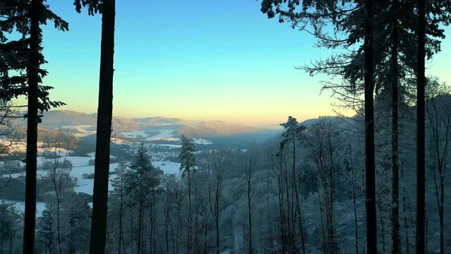 Flight past trees towards snow covered valley and village in winter at sunset