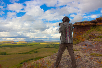 Fototapeta premium A brunet man traveler with a stands on the edge of a cliff and photographs an autumn steppe landscape under cloudy sky. Mountain range Sunduki in Khakassia, Russia. View from the back