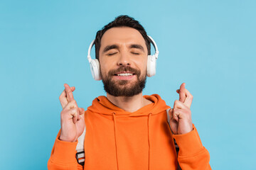 positive student in wireless headphones holding crossed fingers while standing with closed eyes isolated on blue.