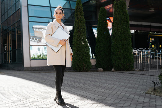 Smiling Young Woman Holding Laptop While Standing In Business District Against Backdrop Of Modern Building Looking Away, Full Length