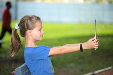 Happy child girl looking at screen of digital tablet resting on bench in park on summer sunny day. Online study concept