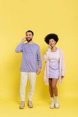 Fototapeta premium full length of smiling interracial couple in stylish clothes talking on smartphones on yellow background.