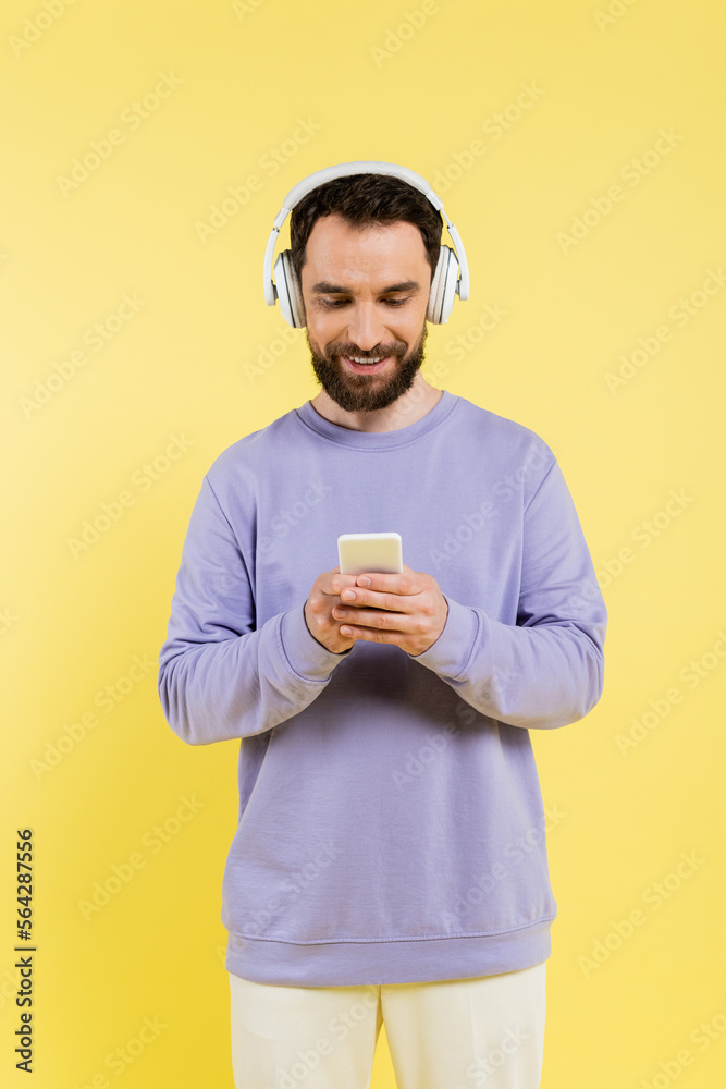 Wall mural smiling bearded man in purple pullover and wireless headphones using smartphone isolated on yellow. - Wall murals