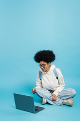 full length of african american student in eyeglasses and white hoodie sitting with crossed legs near laptop on blue background.