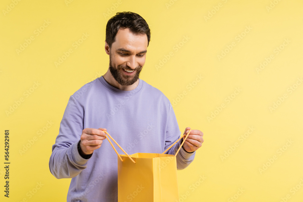 Wall mural pleased bearded man looking into shopping bag isolated on yellow. - Wall murals
