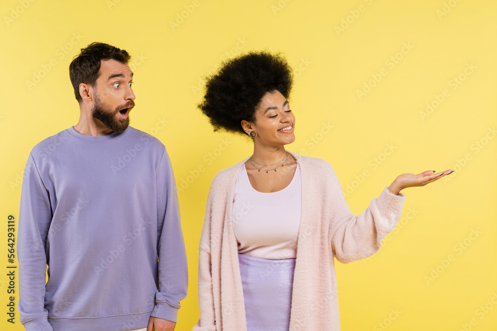 Wall mural smiling african american woman pointing with hand near amazed bearded man isolated on yellow. - Wall murals