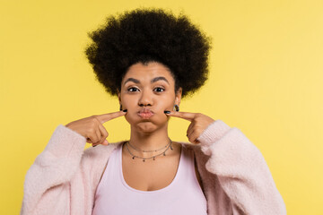 funny african american woman in silver necklaces touching puffed cheeks isolated on yellow.
