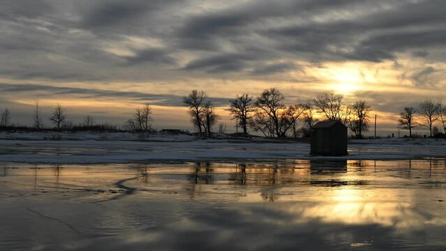 Time lapse of late day sunset clouds moving above an ice fishing shack on a frozen lake. Water on top of the ice is reflecting the clouds.
