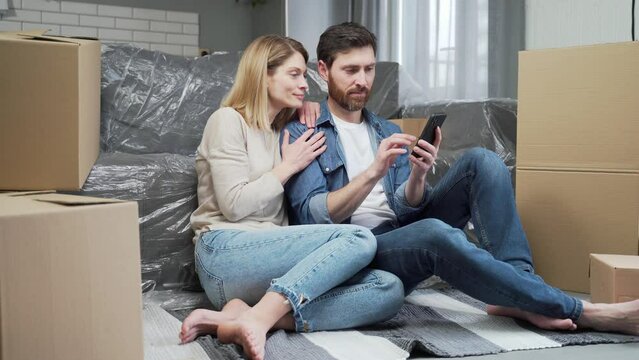 Smiling married couple plan apartment decoration browsing online stores using smartphone sitting in a living room of their new home with a lot of boxes in the background Moving relocation concept
