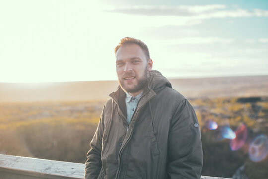 Portrait of attractive man with beard standing on the wooden bridge and looking at camera. Smiling male enjoying the sun